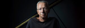 Colour portrait of Keith Jarrett in front of a piano, facing the camera.