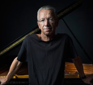 Colour portrait of Keith Jarrett in front of a piano, facing the camera.